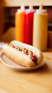 A classic hot dog in front of several bottles of condiments.