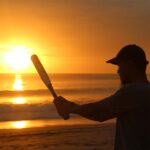 A man swings a bat standing in front of the ocean with the sun setting behind him.