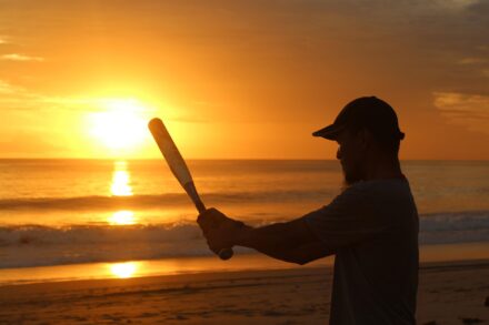 A man swings a bat standing in front of the ocean with the sun setting behind him.