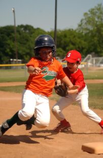 A baseball player is running toward a base, while the player behind him is catching the ball.