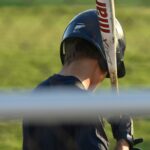 A baseball player holds his bat while looking at the ground.