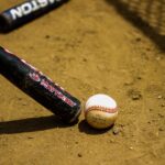 A baseball bat and ball are in the dirt in front of a chain-link fence.