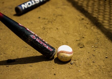 A baseball bat and ball are in the dirt in front of a chain-link fence.