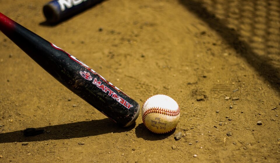 A baseball bat and ball are in the dirt in front of a chain-link fence.