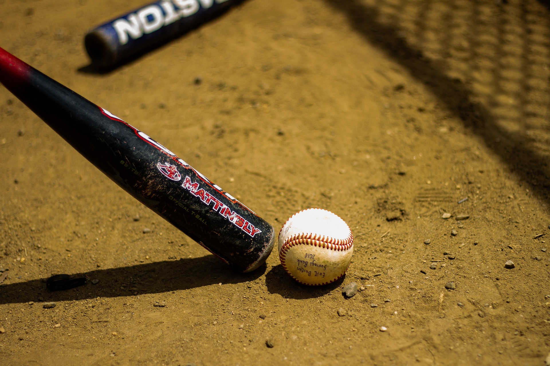 A baseball bat and ball are in the dirt in front of a chain-link fence.