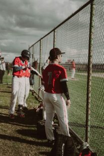 Baseball players watch the game through a chain-link fence.