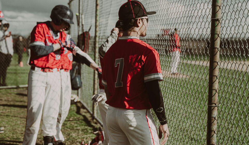 Baseball players watch the game through a chain-link fence.