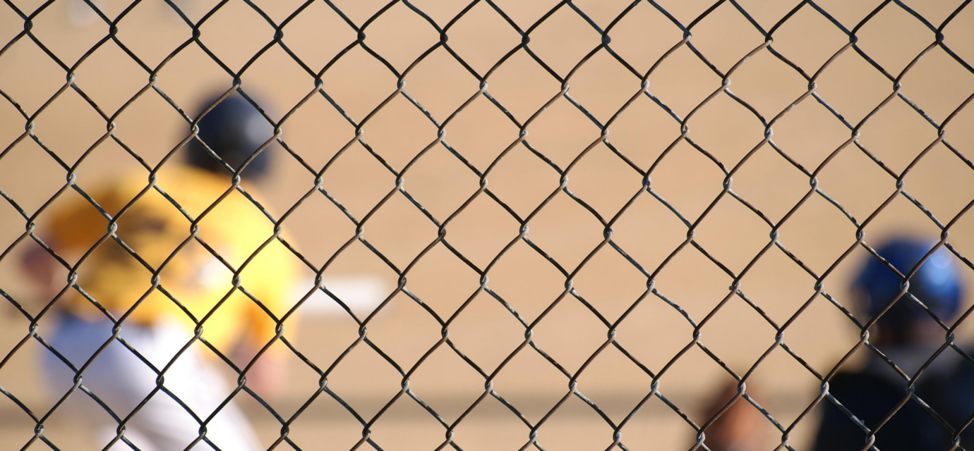 A baseball player can be seen at bat through a chain-link fence.