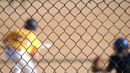 A baseball player can be seen at bat through a chain-link fence.