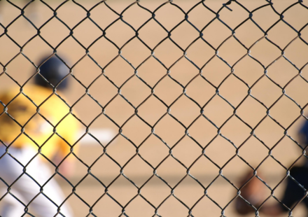 A baseball player can be seen at bat through a chain-link fence.