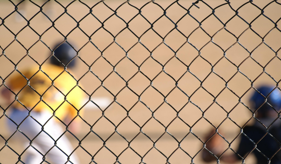 A baseball player can be seen at bat through a chain-link fence.