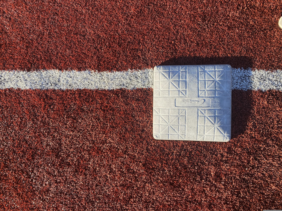 White first base centered on a foul line on a red artificial-turf baseball field.