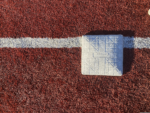 White first base centered on a foul line on a red artificial-turf baseball field.