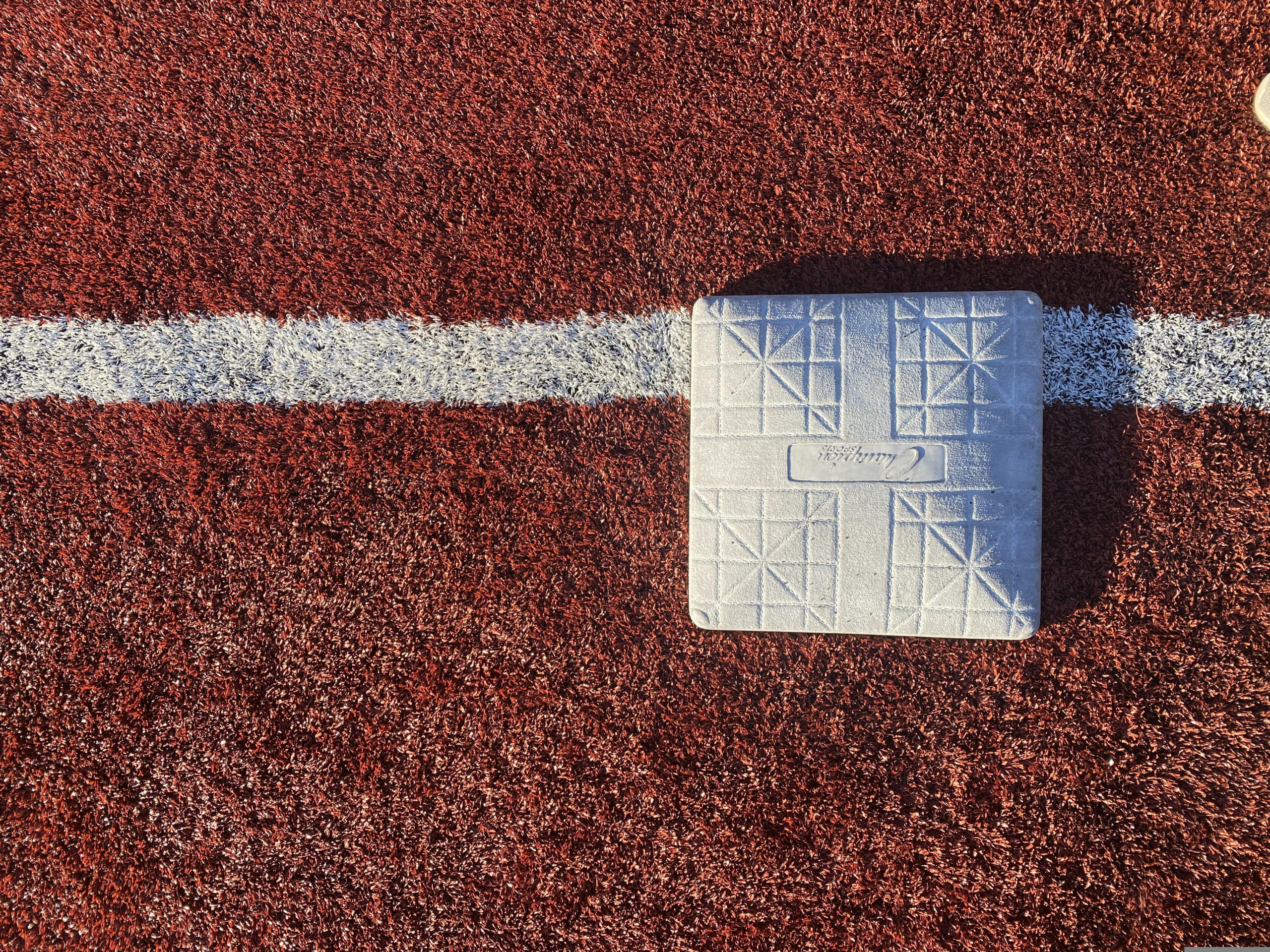 White first base centered on a foul line on a red artificial-turf baseball field.