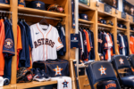 Houston Astros uniforms and equipment fill a brightly lit clubhouse locker beside an MLB Postseason credential.