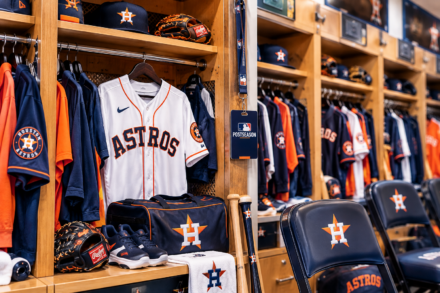 Houston Astros uniforms and equipment fill a brightly lit clubhouse locker beside an MLB Postseason credential.