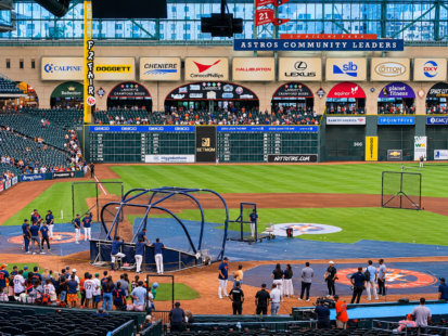Houston Astros players and coaches gather around the batting cage during batting practice at Daikin Park, with the outfield scoreboard and stadium seating in the background.