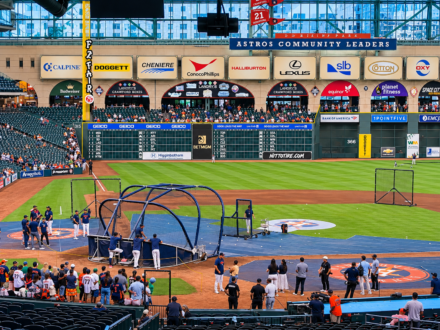 Houston Astros players and coaches gather around the batting cage during batting practice at Daikin Park, with the outfield scoreboard and stadium seating in the background.