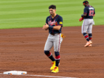 Mauricio Dubón smiles and claps near third base in his Atlanta Braves uniform at Daikin Park, wearing bright yellow cleats, with another Braves player in the background.