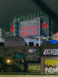 Fenway Park’s illuminated Boston Red Sox sign rises above graffiti-covered buildings at night.