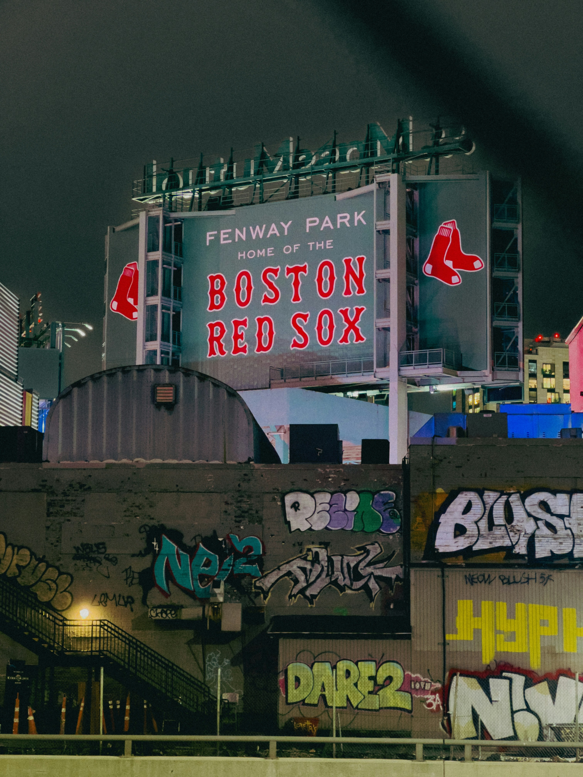 Fenway Park’s illuminated Boston Red Sox sign rises above graffiti-covered buildings at night.