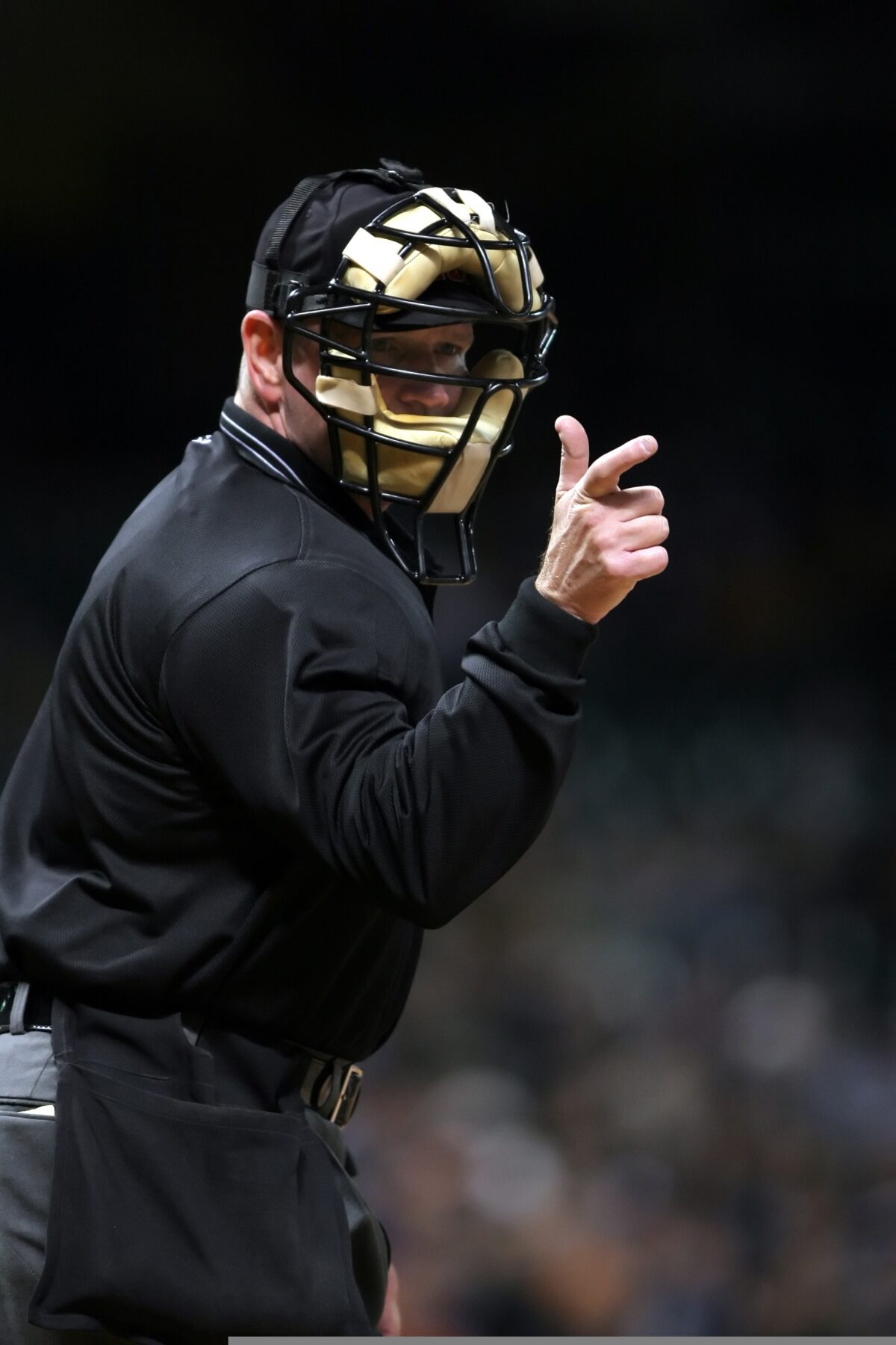 A home plate umpire wearing a protective mask and black uniform points while signaling a call during a baseball game.